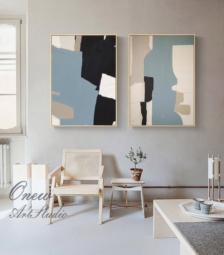 May include: Two abstract paintings with a blue, black, white, and beige color scheme. The paintings are framed in gold and hung on a light gray wall.