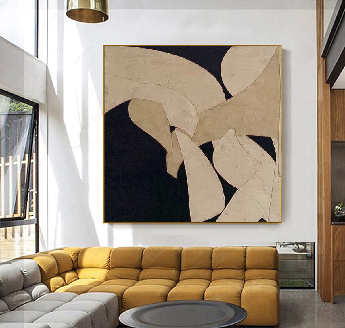 May include: Abstract art painting with a gold frame. The artwork features beige and black shapes. The painting is displayed in a modern living room with a yellow sofa and a gray ottoman.
