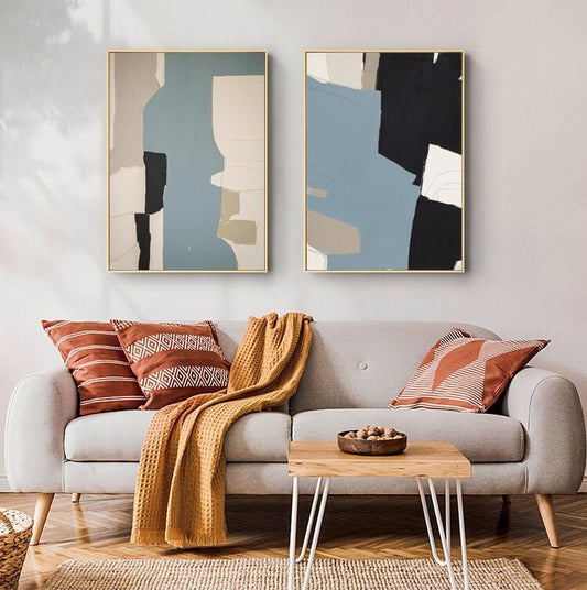 May include: Two abstract paintings with geometric shapes in shades of blue, black, white, and beige. The paintings are framed in gold and hung on a white wall above a gray sofa.
