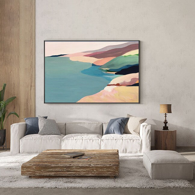 May include: Abstract landscape painting with a coastal scene featuring a blue body of water, sandy beaches, and colorful hills in shades of pink, green, and brown. The painting is framed in a black frame and is hanging on a white wall.