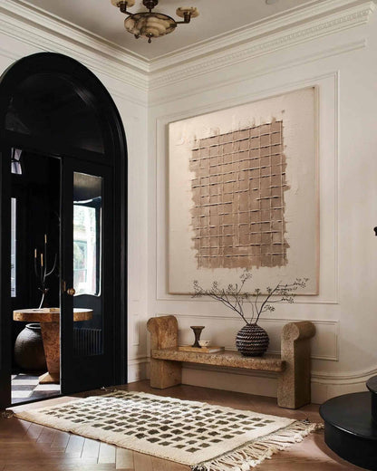 May include: A large abstract painting with a grid pattern in shades of brown and beige hangs on a white wall above a stone bench. The bench has a vase with dried branches and a small ceramic vase on top. The room has a black door with a white and black checkered floor.