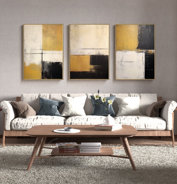 May include: Three abstract paintings in gold, white, and black frames. The paintings are in a minimalist style with geometric shapes and muted colors. The paintings are hung on a light gray wall above a white sofa with pillows.