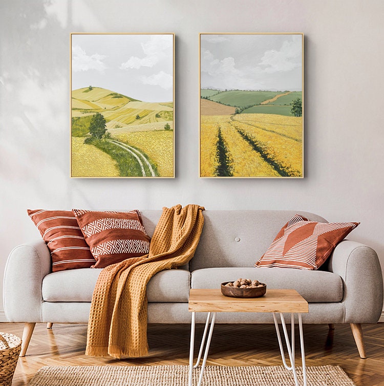 May include: Two framed landscape paintings depicting rolling hills and fields of yellow wildflowers. The paintings are hung on a white wall above a gray sofa with throw pillows and a blanket.