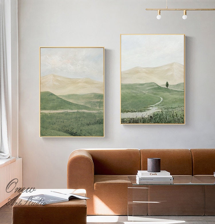 May include: Two framed landscape paintings with a green and brown color scheme. The paintings depict rolling hills, a winding path, and a lone tree. The paintings are hung on a white wall above a brown couch.