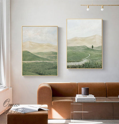 May include: Two framed landscape paintings with a green and brown color scheme. The paintings depict rolling hills, a winding path, and a lone tree. The paintings are hung on a white wall above a brown couch.
