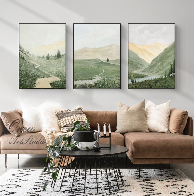 May include: Three framed landscape paintings with a green and brown color scheme depicting rolling hills, a winding path, and a small lake. The paintings are hung on a white wall above a brown sectional sofa with pillows and a black coffee table.