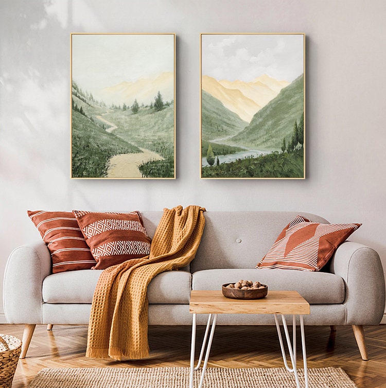 May include: Two framed landscape paintings of a mountain valley with a winding path and a lake. The paintings are in a living room setting with a couch, throw pillows, and a coffee table.