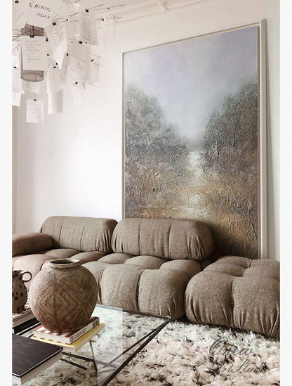 May include: A large abstract painting of a landscape with a path leading to a misty horizon. The painting is framed in a white frame and hangs above a brown sectional sofa. The sofa is in a living room with a glass coffee table and a large brown vase.