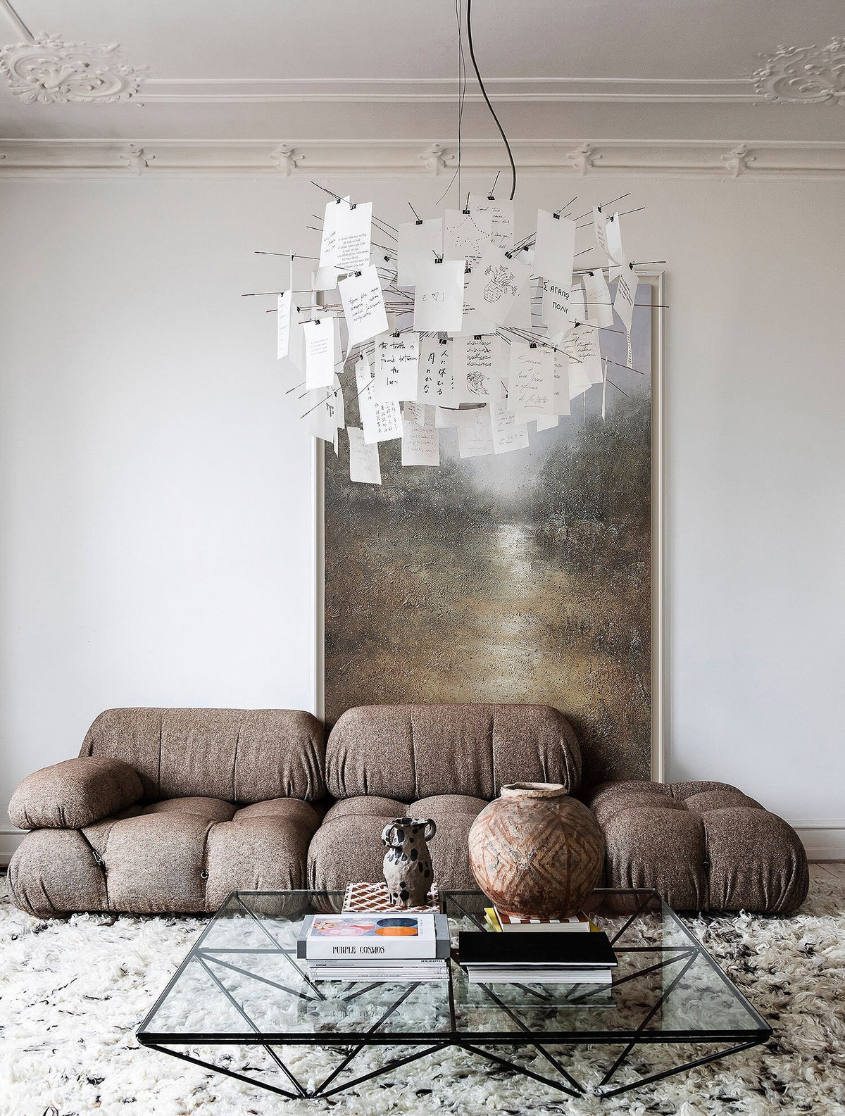 May include: A brown sectional sofa with a glass coffee table in front of it. The coffee table has a black metal frame and is decorated with books and a ceramic vase. A large abstract painting hangs on the wall behind the sofa.