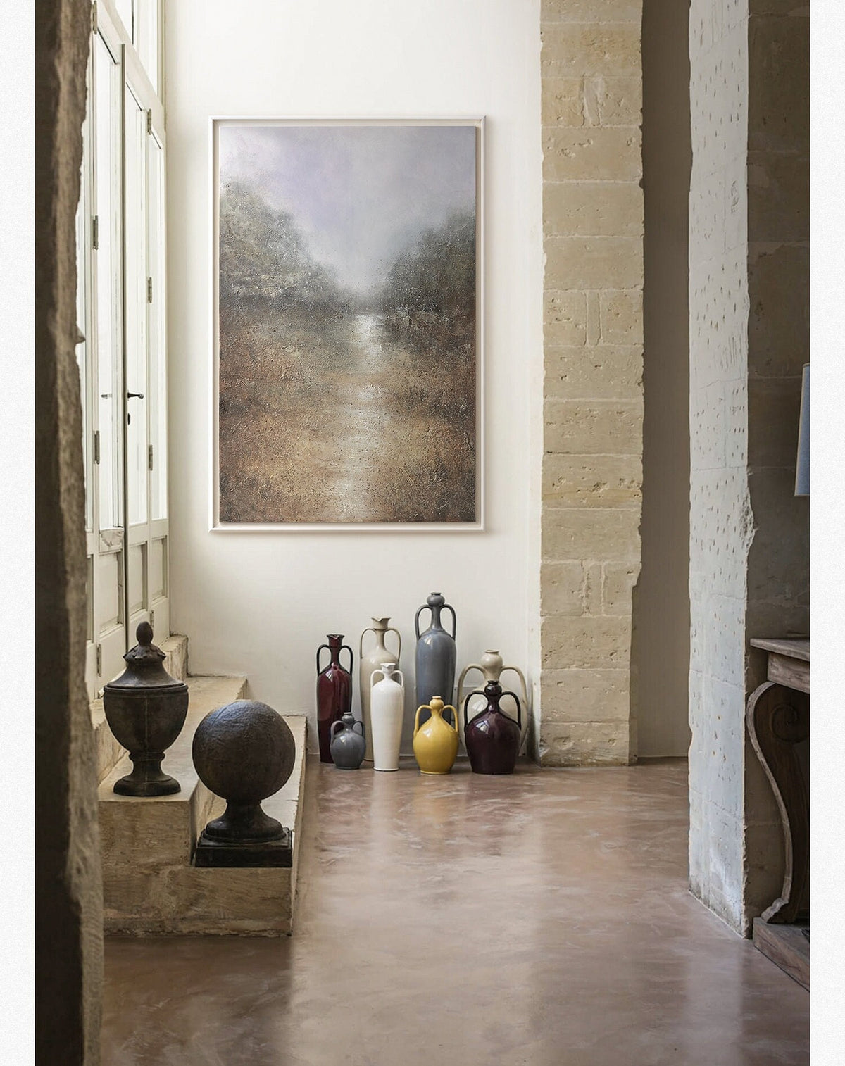 May include: A collection of ceramic vases in various colors and styles, including a white vase, a yellow vase, a gray vase, a brown vase, and a red vase. The vases are arranged on a light brown floor in front of a white wall with a framed painting of a landscape.