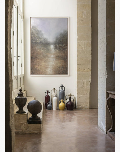 May include: A collection of ceramic vases in various colors and styles, including a white vase, a yellow vase, a gray vase, a brown vase, and a red vase. The vases are arranged on a light brown floor in front of a white wall with a framed painting of a landscape.