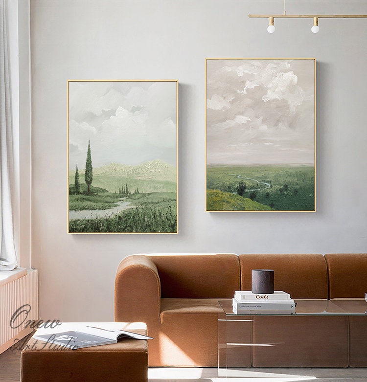 May include: Two framed landscape paintings with a green and brown color scheme. The painting on the left depicts a green field with a stream and trees. The painting on the right depicts a green field with a winding road and a cloudy sky.