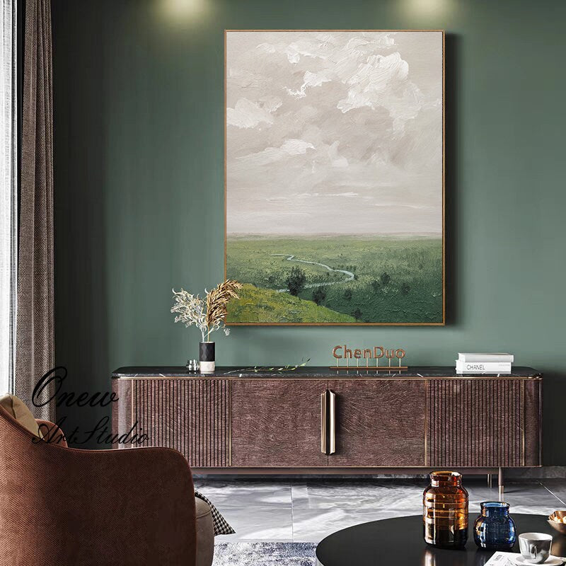 May include: A large abstract landscape painting with a green field, a winding river, and a cloudy sky. The painting is framed and hanging on a green wall above a brown wooden cabinet.