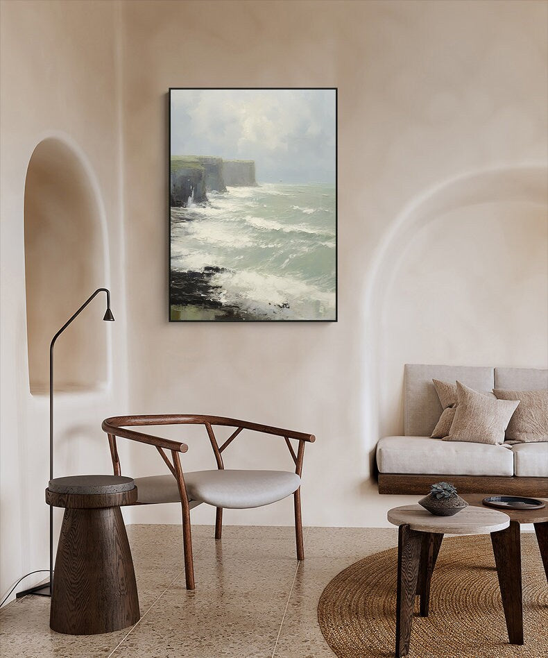 May include: A framed painting of a seascape with a rocky cliff and crashing waves. The painting is in a minimalist living room setting with a chair, a side table, and a sofa.