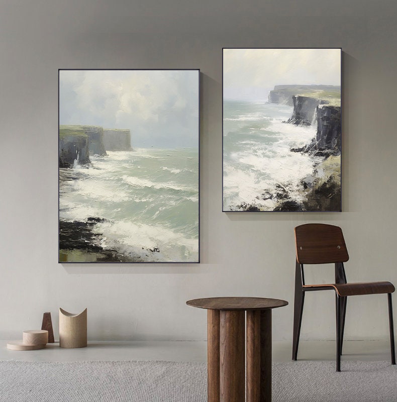 May include: Two framed paintings of seascapes with rocky cliffs and crashing waves. The paintings are hung on a light gray wall with a wooden table and chair in the foreground.