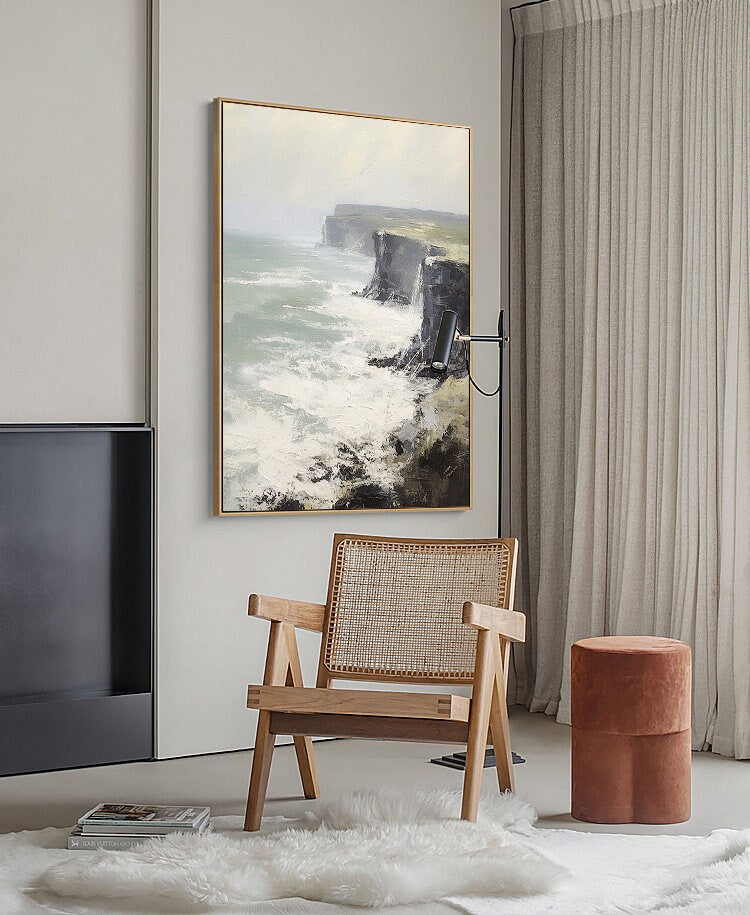 May include: A framed painting of a seascape with a rocky cliff and crashing waves. The painting is hanging on a white wall in a room with a wooden chair and a brown ottoman.
