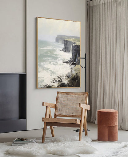 May include: A framed painting of a seascape with a rocky cliff and crashing waves. The painting is hanging on a white wall in a room with a wooden chair and a brown ottoman.