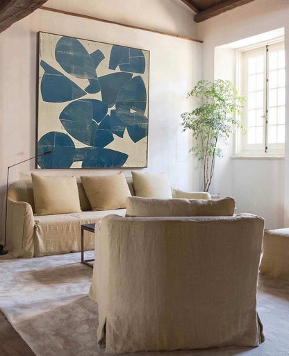 May include: A living room scene featuring a large abstract painting with blue shapes on a white background. A beige sofa with matching pillows and two armchairs are in front of the painting. A small plant sits near a window.