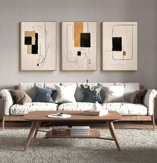 May include: Three abstract art prints with black, white, and orange geometric shapes and lines on a white background. The prints are framed in gold and hung on a white wall above a white sofa.