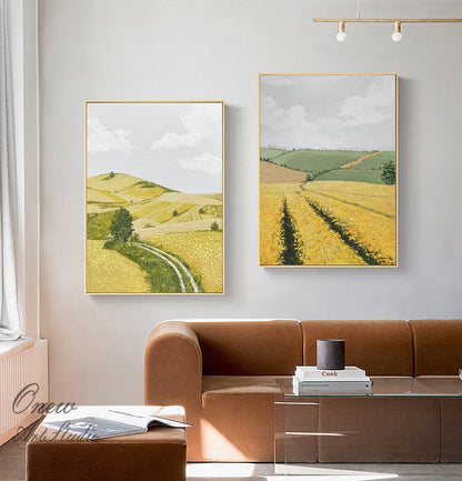 May include: Two framed landscape paintings depicting rolling hills and fields of yellow wildflowers. The paintings are hung on a white wall above a brown sofa.