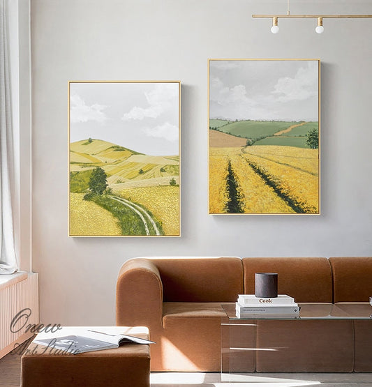 May include: Two framed landscape paintings depicting rolling hills and fields of yellow wildflowers. The paintings are hung on a white wall above a brown sofa.