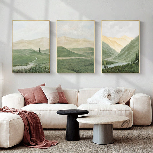 May include: Three framed landscape paintings with a green and brown color scheme. The paintings depict rolling hills, a river, and a single tree in a field. The paintings are hung on a white wall above a white couch with two throw pillows.