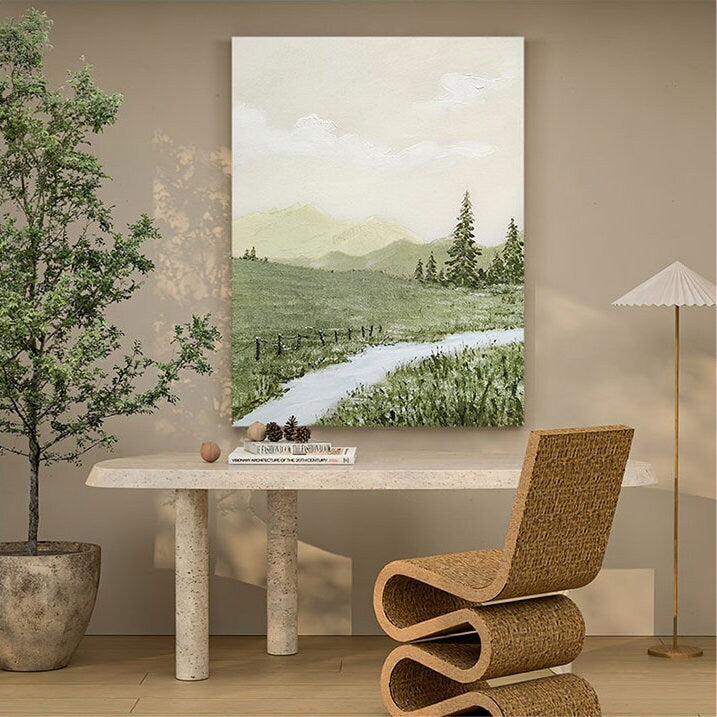 May include: A painting of a green landscape with a winding path leading to a mountain range in the distance. The painting is framed and hanging on a wall above a white table with a book and a chair.