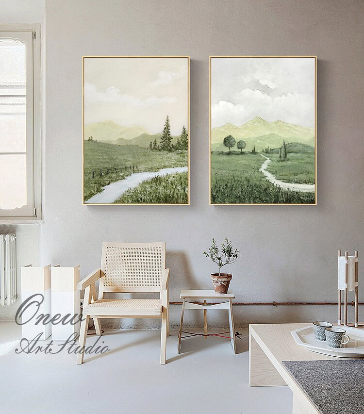 May include: Two framed landscape paintings with green and white hues. The paintings depict a winding path through a grassy field with mountains in the background. One painting features a stream running through the field.