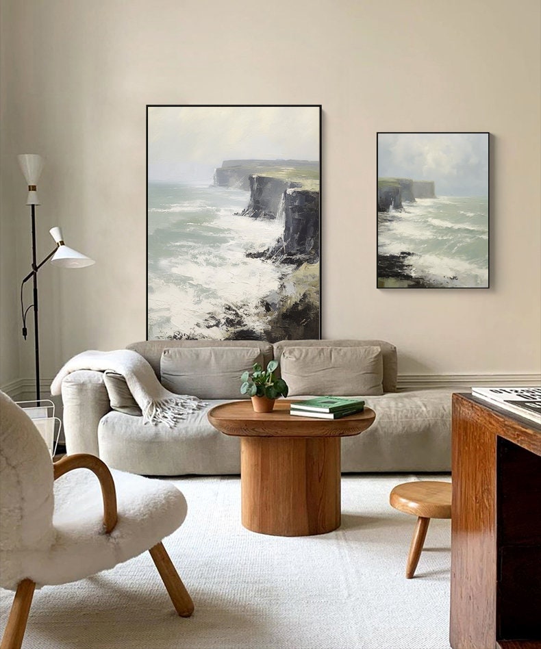 May include: Two framed paintings of a seascape with crashing waves and cliffs. The paintings are hanging on a light beige wall above a light gray couch. A wooden coffee table with a potted plant and books sits in front of the couch.