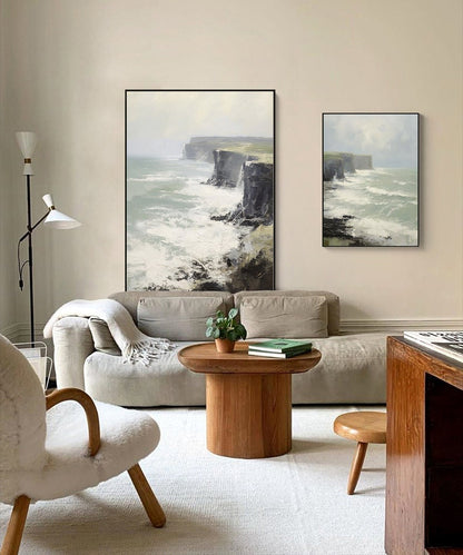 May include: Two framed paintings of a seascape with crashing waves and cliffs. The paintings are hanging on a light beige wall above a light gray couch. A wooden coffee table with a potted plant and books sits in front of the couch.