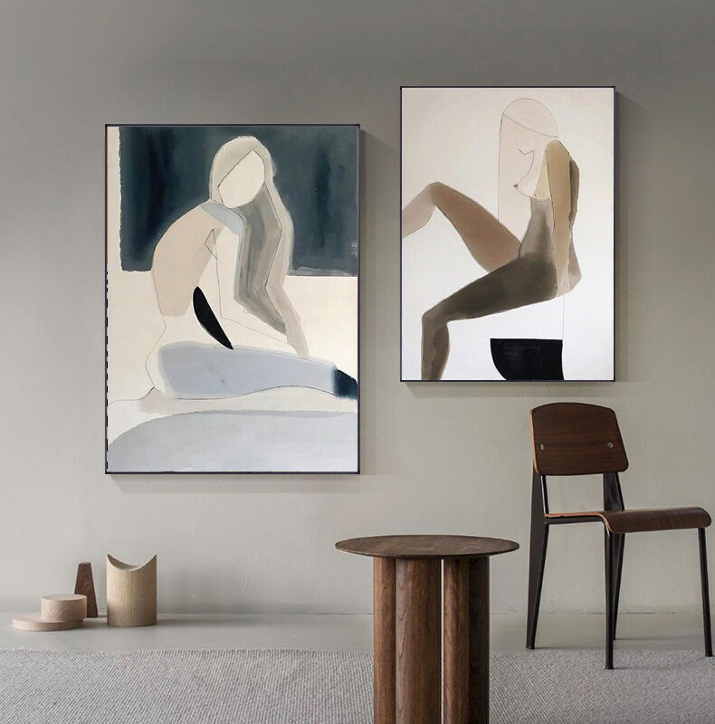 May include: Two abstract paintings of figures in muted tones of brown, gray, and white. The paintings are framed in black and hung on a light gray wall.