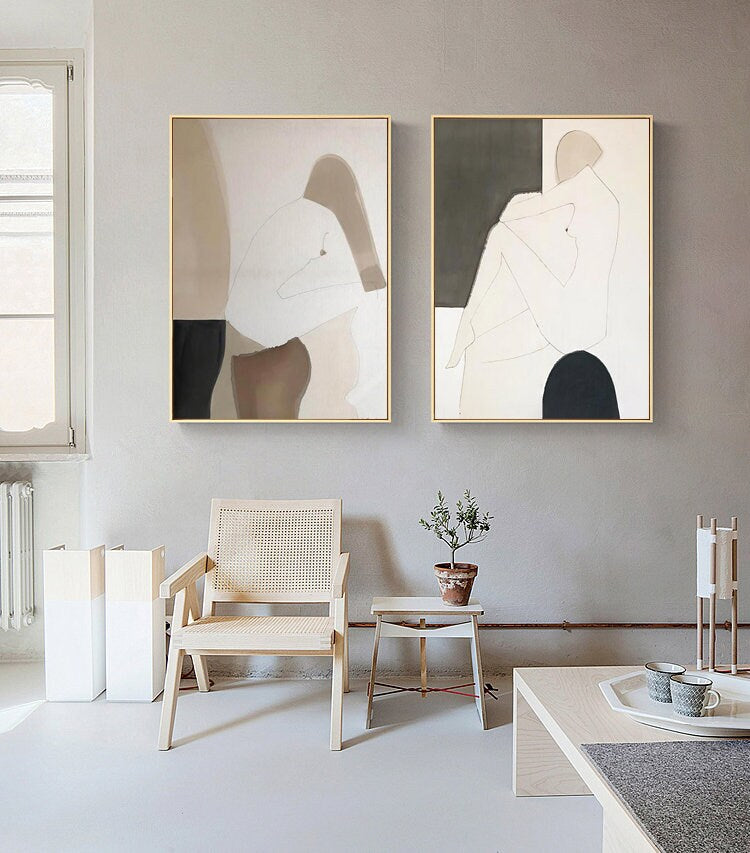May include: Two abstract paintings with a minimalist style featuring nude figures in muted tones of brown, beige, and black. The paintings are framed in gold and hung on a light gray wall.