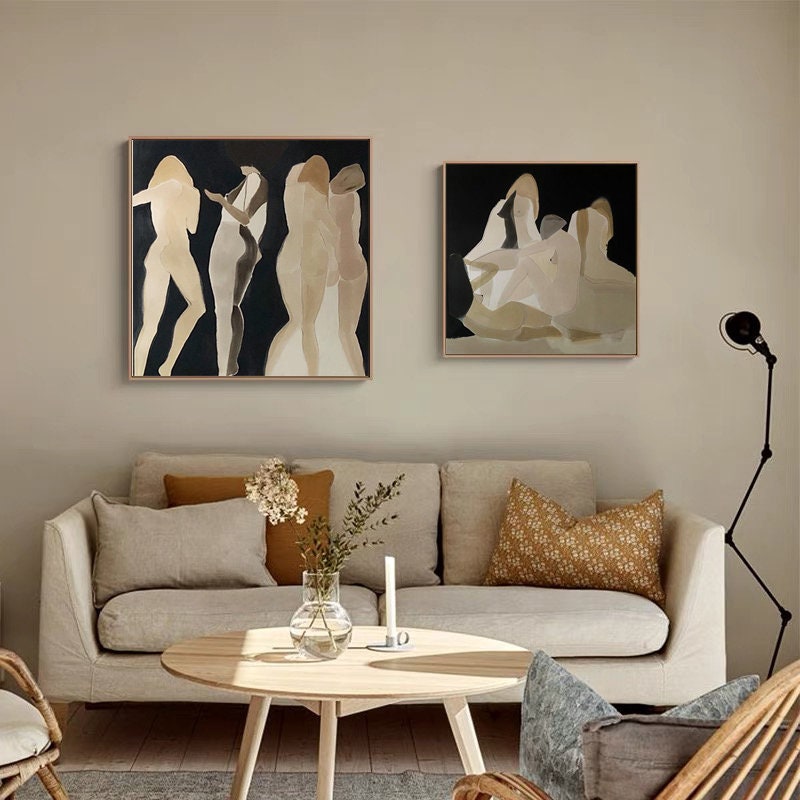 May include: Two abstract paintings of figures in muted tones on a black background. The paintings are framed and hung on a beige wall above a light beige sofa with brown throw pillows.