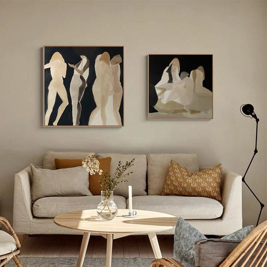 May include: Two abstract paintings of figures in muted tones on a black background. The paintings are framed and hung on a beige wall above a light beige sofa with brown throw pillows.