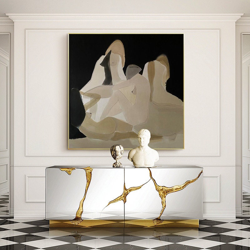 May include: A gold and silver console table with a unique, abstract design. The table has two doors and is decorated with gold accents. The table is in front of a white wall with a large abstract painting of figures in shades of brown and black.