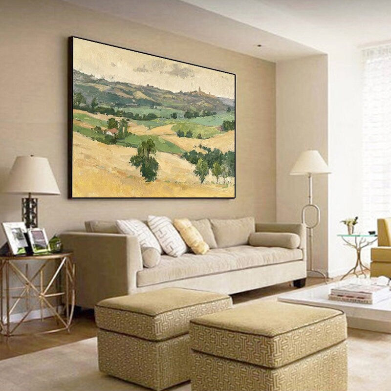 May include: A large framed landscape painting depicting rolling hills and fields in shades of green, yellow, and beige. The artwork hangs above a beige sofa with decorative pillows. The room also features two matching ottomans and a side table.