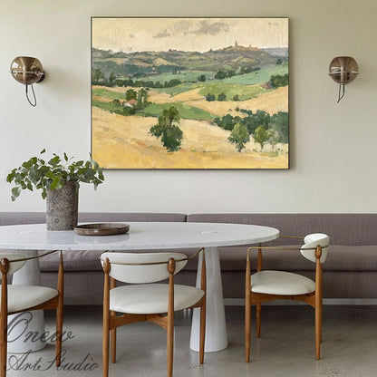 May include: An oil painting of a rural landscape with rolling hills, fields, and trees. The artwork features a palette of yellow, green, and beige hues, with a distant town or building visible on a hilltop. The painting is framed and hangs above a dining table.