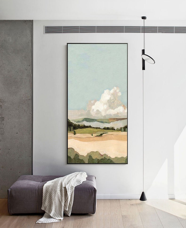 May include: A framed abstract landscape painting with a light blue sky, white clouds, and green and brown hills. The painting is hanging on a white wall in a modern living room.