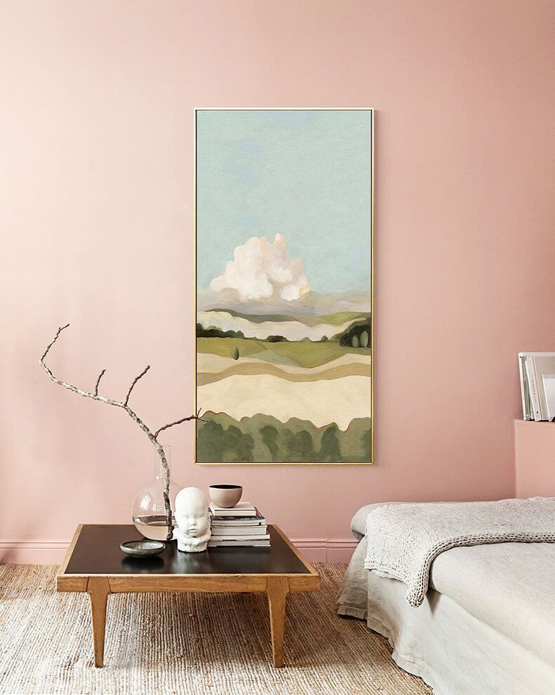 May include: A vertical landscape painting with a light blue sky, a large white cloud, and rolling green hills. The painting is framed in a gold frame and is hanging on a light pink wall.