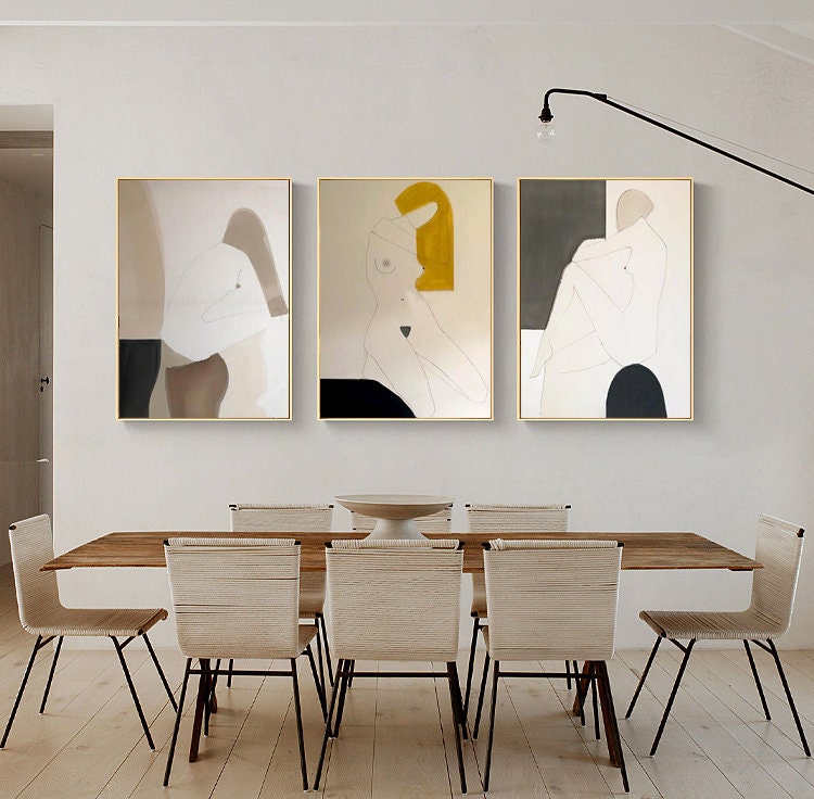 May include: Three abstract art prints with gold frames, featuring minimalist line drawings of figures in various poses. The prints are hung on a white wall above a wooden dining table with chairs.