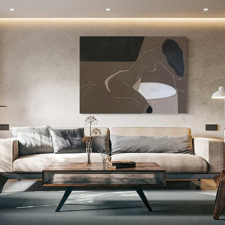 May include: A beige sofa with gray pillows in a living room setting. A painting of a nude figure in a minimalist style hangs on the wall above the sofa. A wooden coffee table sits in front of the sofa.