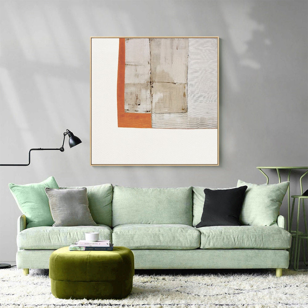 May include: A green sofa with three pillows, a green ottoman, and a framed abstract painting with orange, white, and brown geometric shapes.