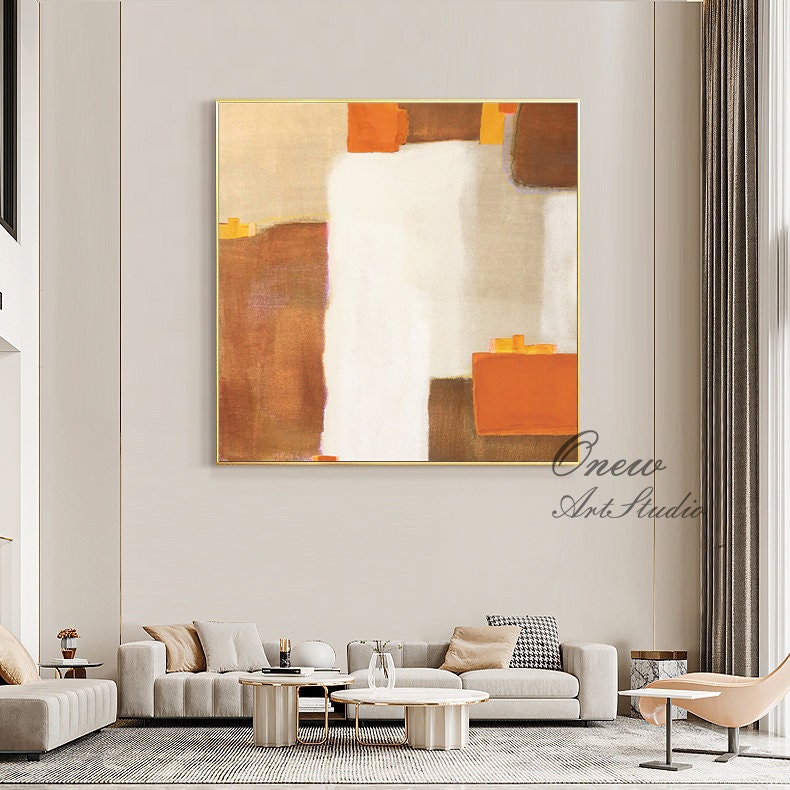 May include: Abstract painting with brown, orange, and white geometric shapes. The painting is framed in gold and hangs on a wall in a living room setting.