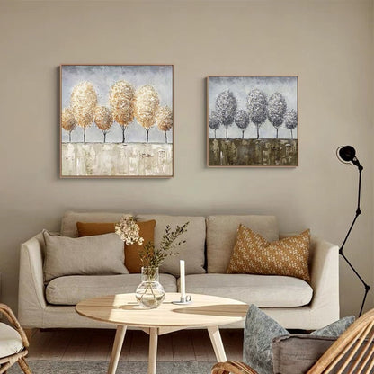 May include: Two square canvas paintings with abstract tree designs. The painting on the left features trees with gold and brown tones, while the painting on the right features trees with gray and white tones. Both paintings are displayed on a wall above a beige sofa with pillows.