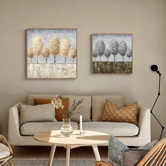 May include: Two square canvas paintings with abstract tree designs. The painting on the left features trees with gold and brown tones, while the painting on the right features trees with gray and white tones. Both paintings are displayed on a wall above a beige sofa with pillows.