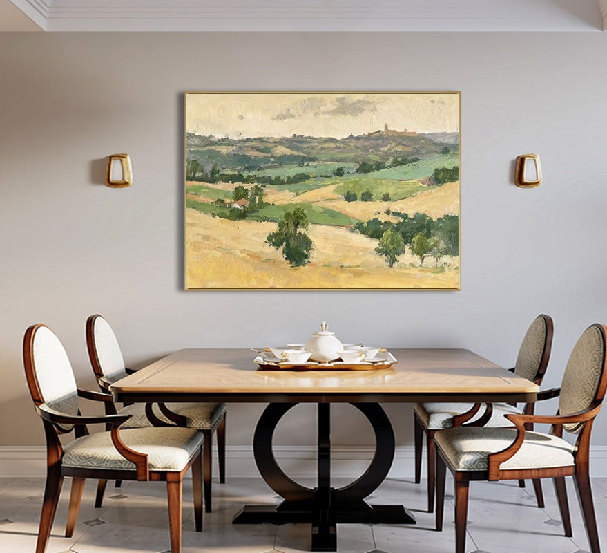 May include: A framed landscape painting depicts rolling hills and fields in shades of green and gold under a cloudy sky. The painting hangs above a wooden dining table with four chairs. A tea set sits on the table.