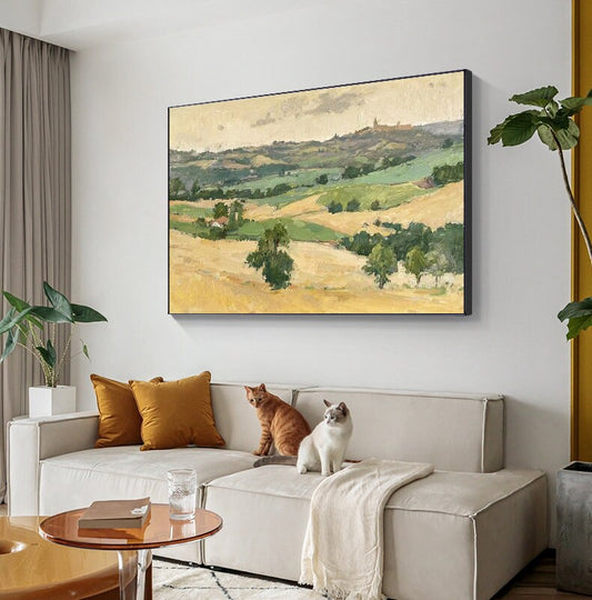 May include: Oil painting of a landscape with rolling hills in shades of green and yellow, under a cloudy sky. The artwork is framed in black and hangs above a cream-colored sofa with two orange throw pillows. Two cats are sitting on the sofa.