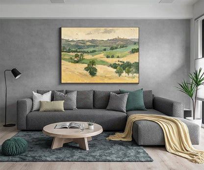 May include: A framed landscape painting depicts rolling hills and fields in shades of green and yellow, under a cloudy sky. The artwork hangs above a gray sectional sofa with decorative pillows, a round wooden coffee table, and a green rug.
