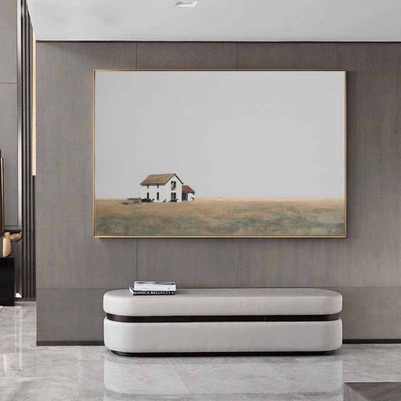 May include: A minimalist painting of a white farmhouse with a brown roof in a field of tall grass. The painting is framed in a gold frame and hangs on a gray wall.