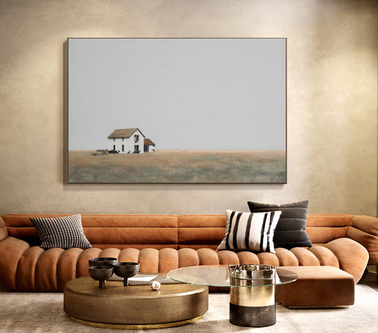 May include: A minimalist painting of a white farmhouse with a brown roof in a field. The painting is framed and hanging on a wall above a brown leather sofa with pillows.